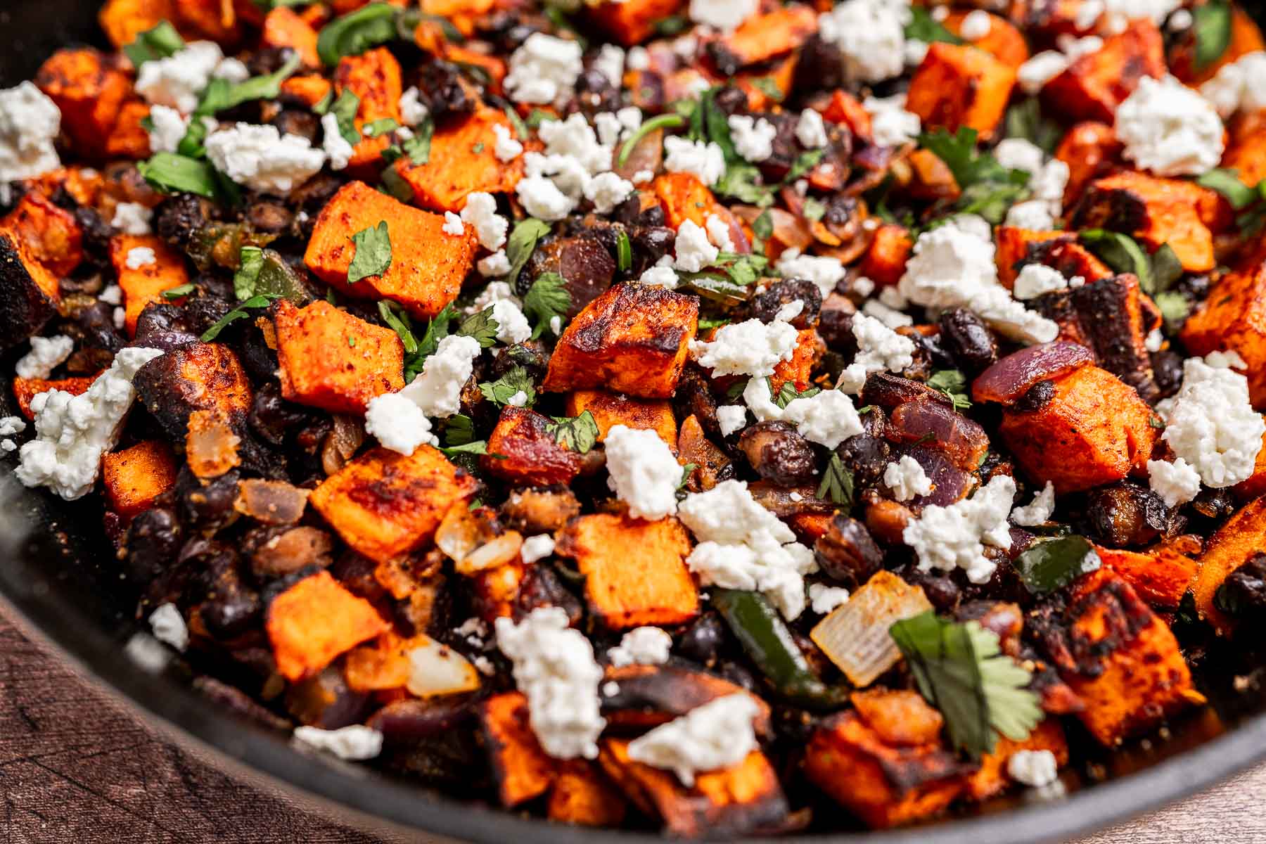 A close-up of a vibrant sweet potato hash featuring roasted sweet potato cubes, black beans, chopped herbs, and crumbled white cheese.