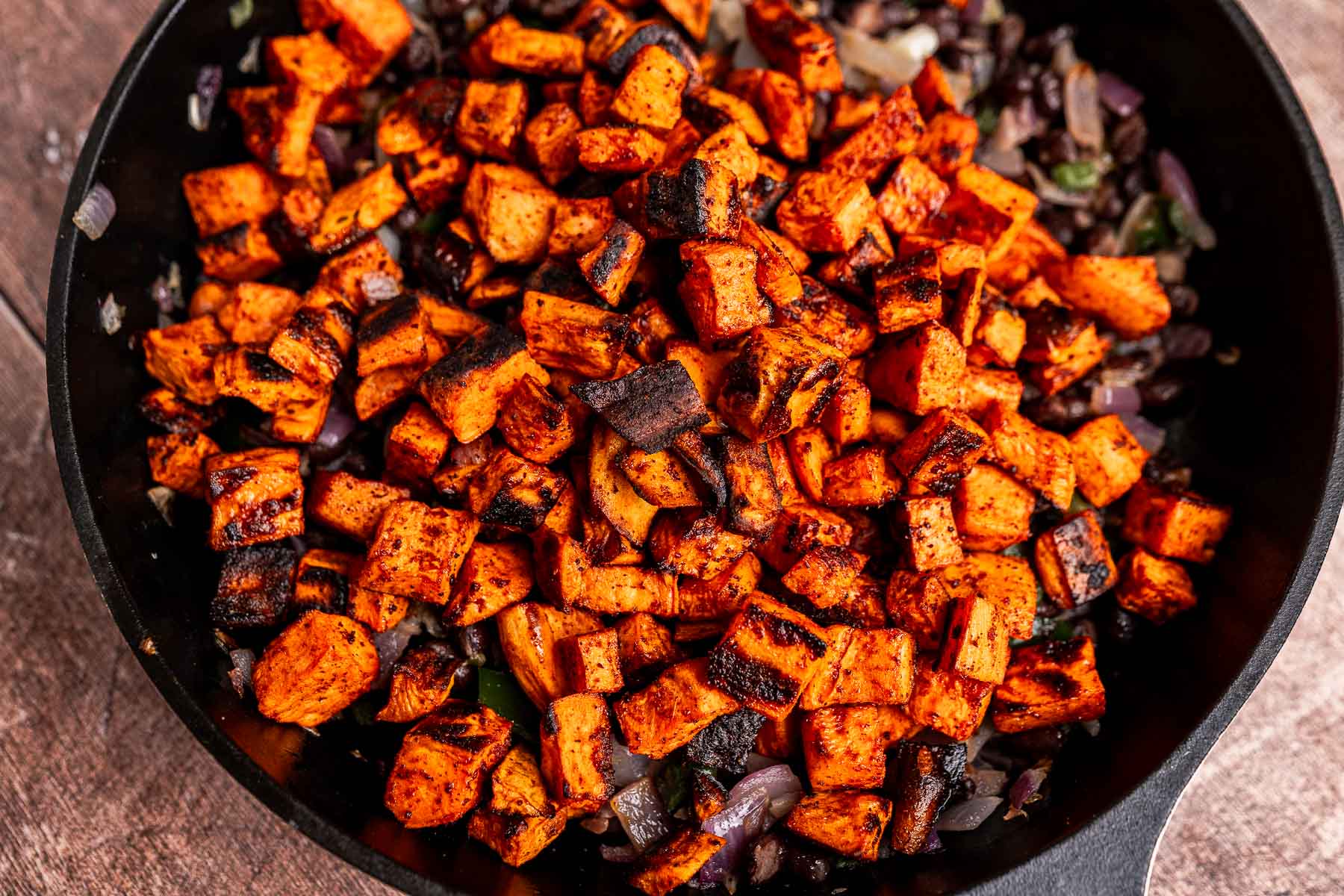 A cast iron skillet filled with sweet potato hash—diced, roasted sweet potatoes and blackened onions—rests on a wooden surface.