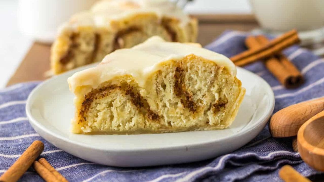A slice of cinnamon roll pie with icing on a white plate, placed on a striped cloth. Cinnamon sticks are nearby.
