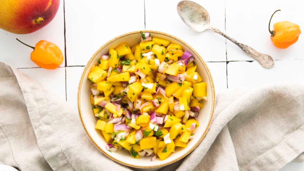 Overhead shot of a bowl of mango habanero salsa with a hand lifting a spoonful of the salsa.