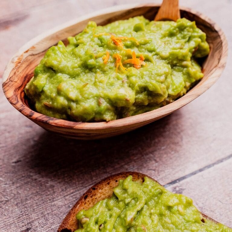 A wooden bowl filled with avocado spread guacamole, topped with orange zest, sits on a wooden surface next to a piece of bread slathered with the creamy avocado spread.