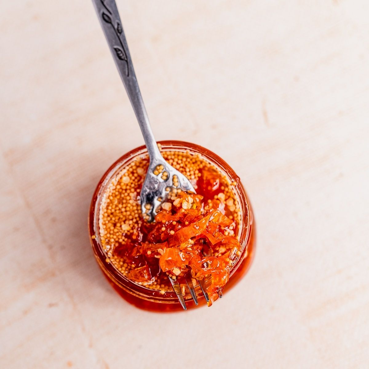 A jar of chili oil with visible seeds, red chili pieces, and candied habaneros, with a fork resting inside the jar, photographed from above on a light surface.