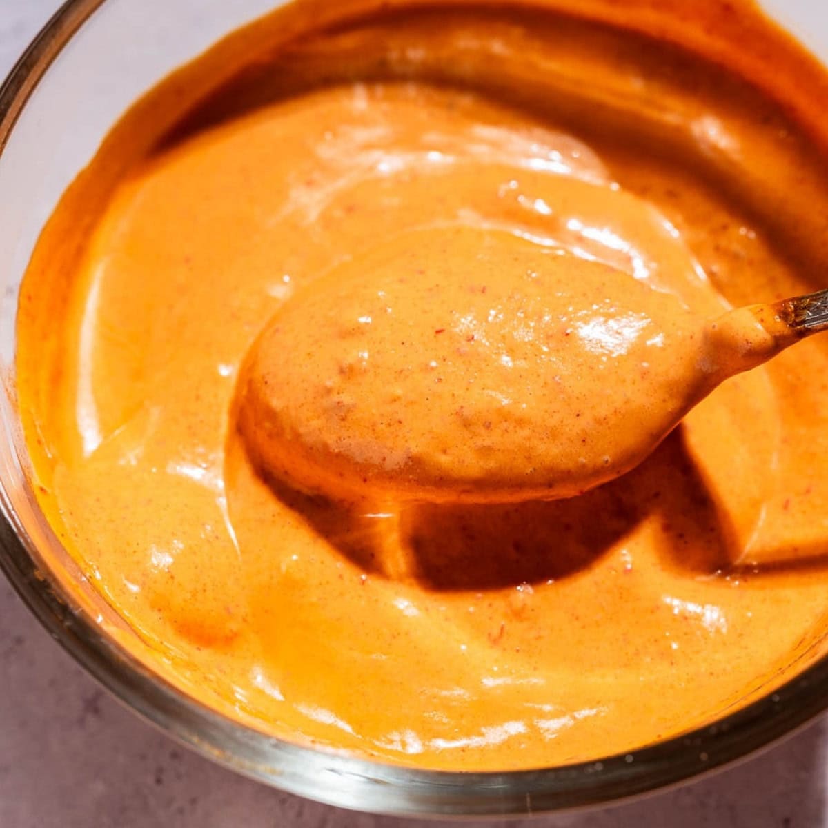 A spoon scooping thick, orange-colored chipotle sauce from a glass bowl on a light surface.