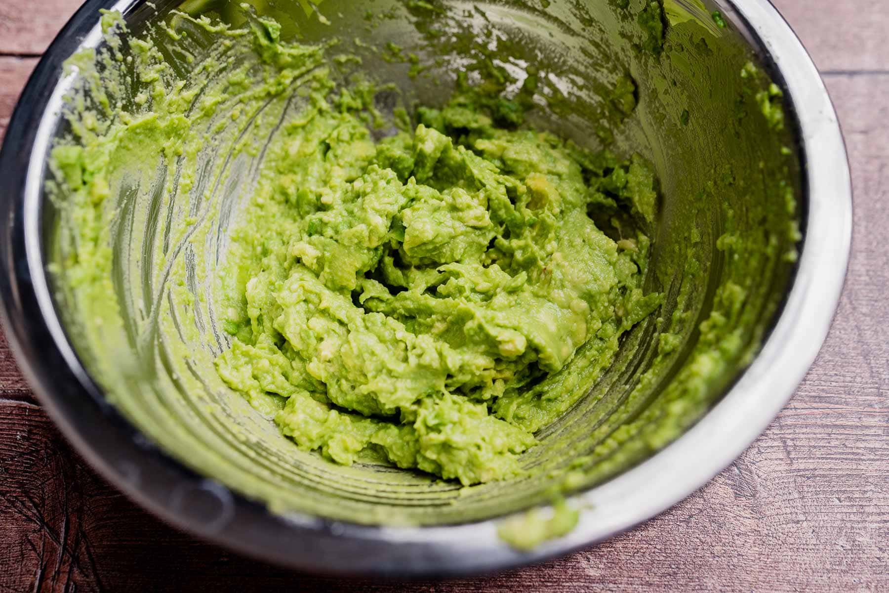 A metal bowl containing freshly mashed avocado spread sits on a wooden surface, with remnants still clinging to the sides.