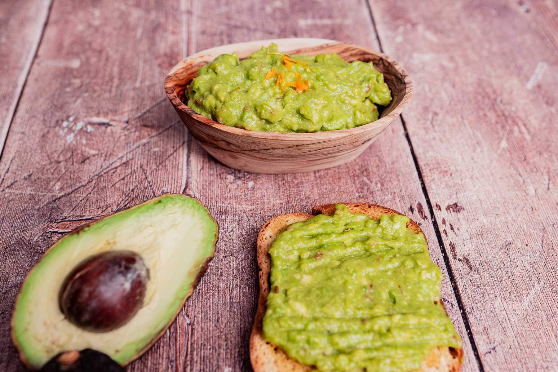A bowl of guacamole, a halved avocado, and a slice of toast topped with creamy avocado spread sit on a wooden surface.