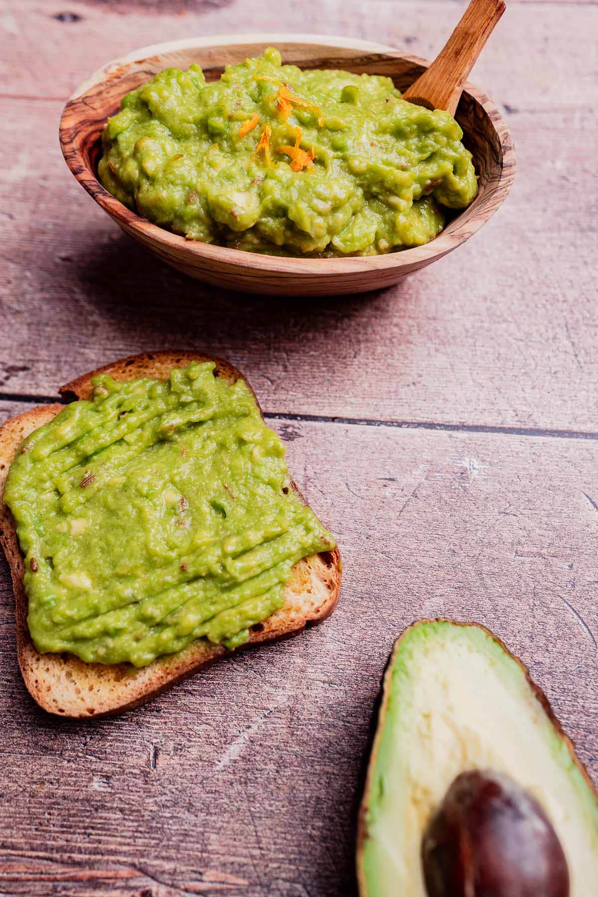 A bowl of guacamole with a wooden spoon, a slice of toast topped with creamy avocado spread, and half an avocado on a wooden surface.