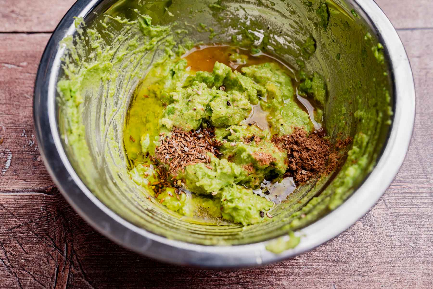 A metal bowl containing avocado spread made with mashed avocado, oil, cumin seeds, and ground spices, placed on a wooden surface.