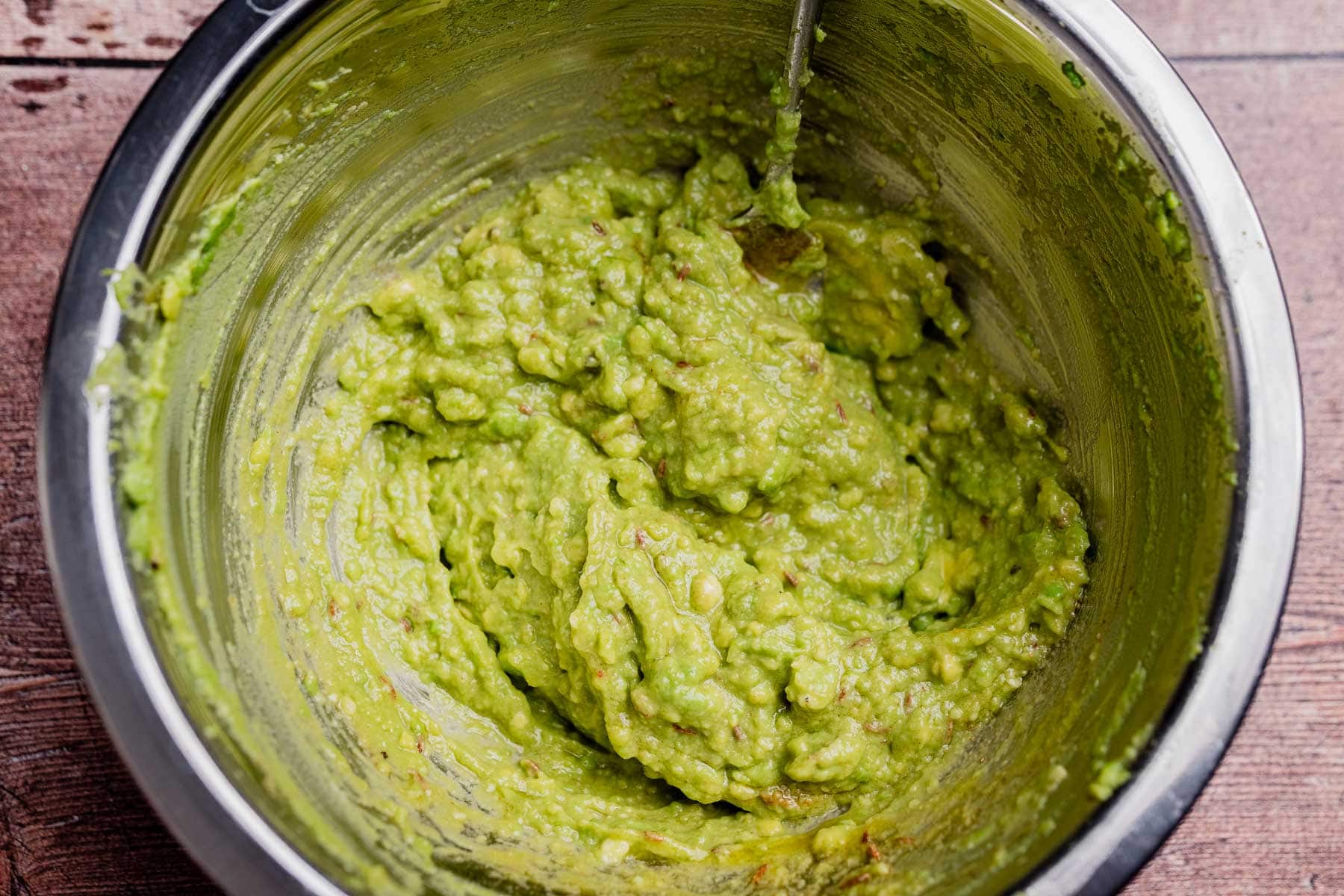 A metal bowl filled with freshly made avocado spread guacamole, with a spoon partially submerged in the mixture.
