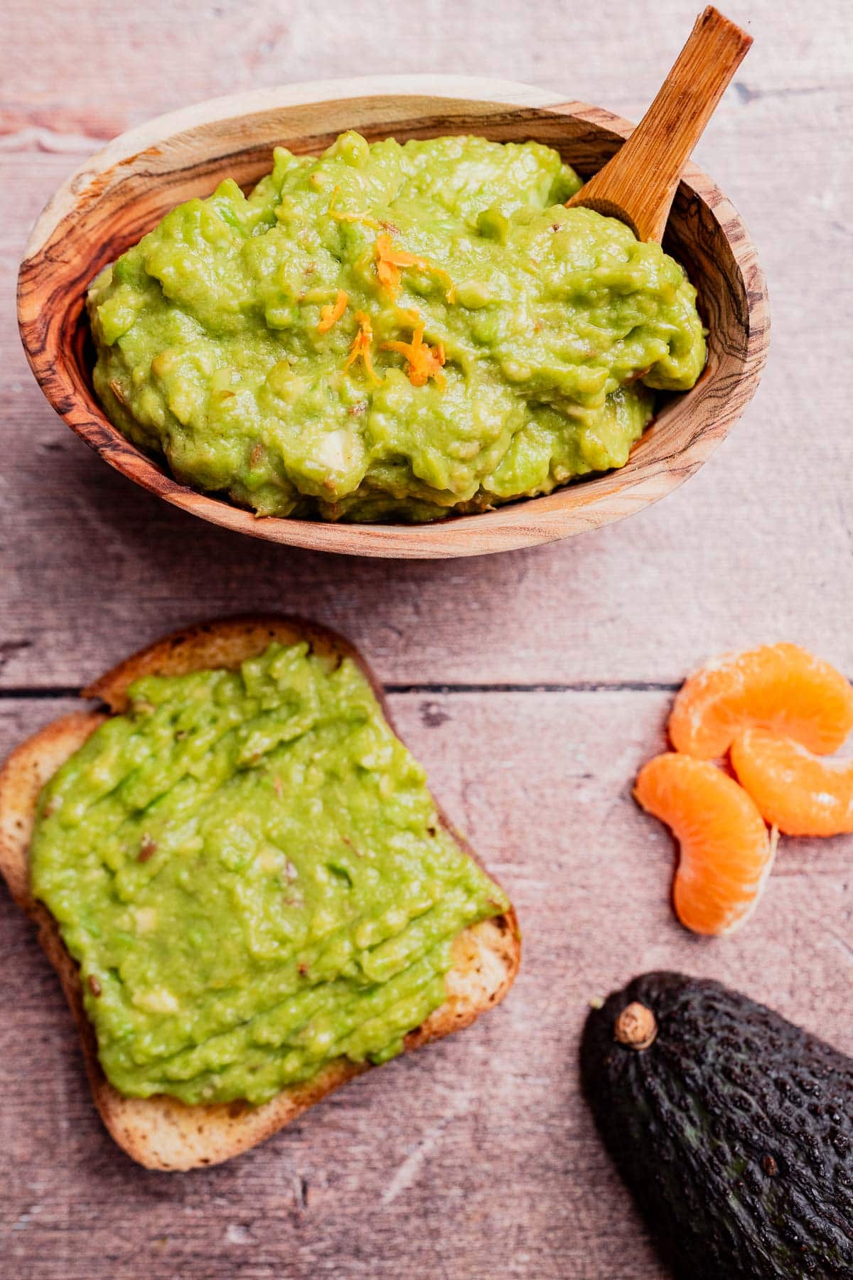 A wooden bowl of avocado spread with a spoon, a slice of toast topped with guacamole, a halved avocado, and tangerine segments on a wooden surface.