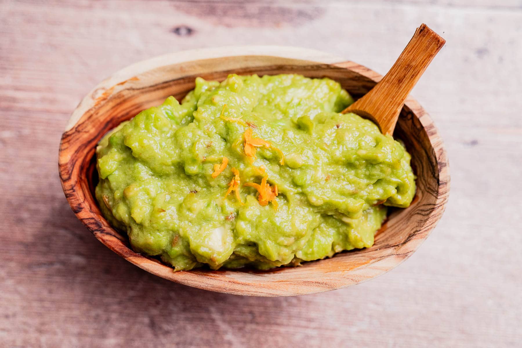 A wooden bowl filled with creamy avocado spread guacamole, topped with a hint of orange zest, with a wooden spoon resting inside.