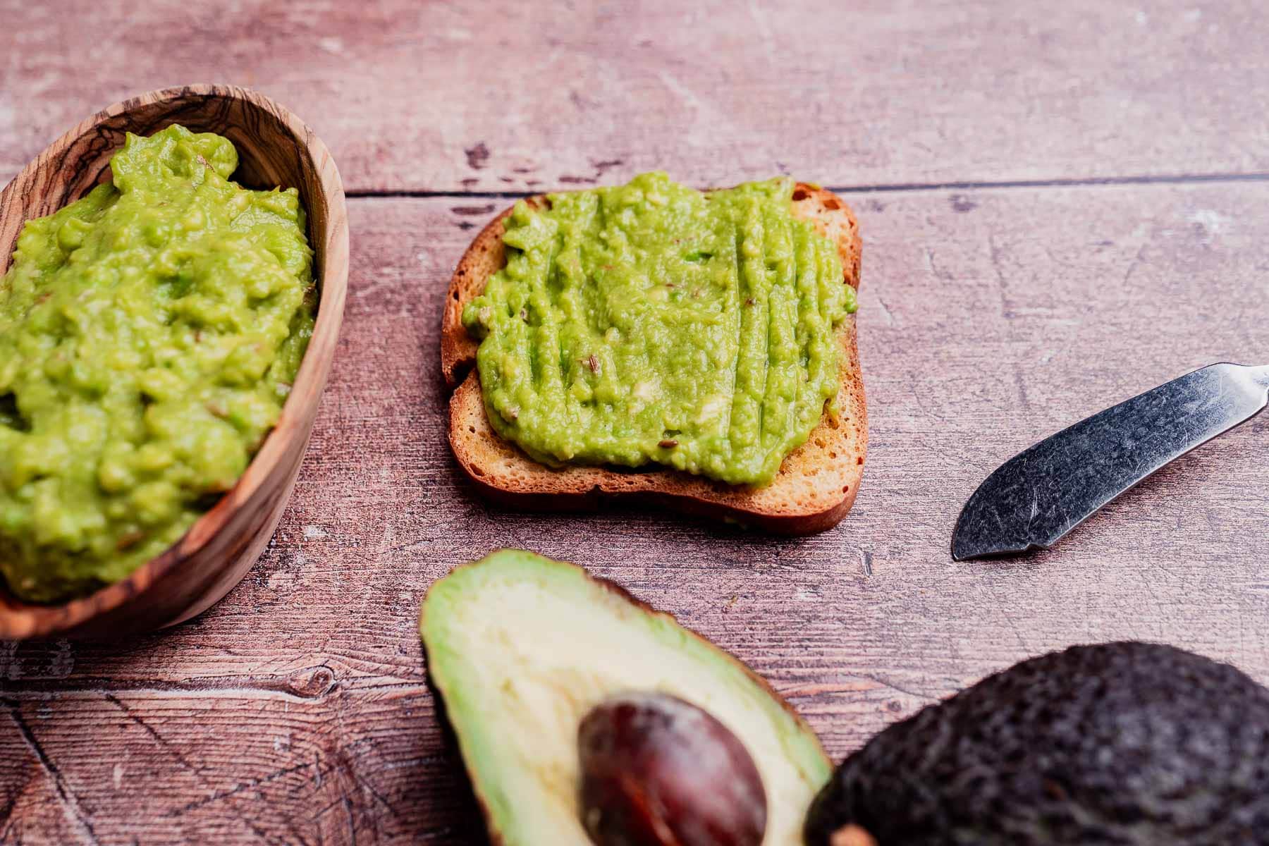 A slice of toast with creamy avocado spread, a bowl of guacamole, a halved avocado with pit, and a knife on a wooden surface.