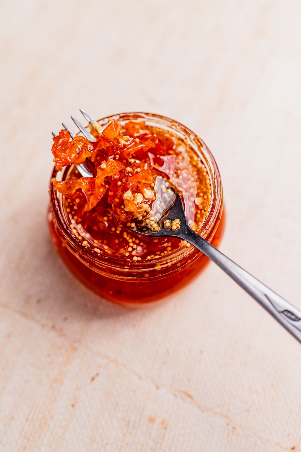 A fork lifts a portion of chili crisp with candied habaneros from an open glass jar filled with spicy oil, chili flakes, and seeds, set on a light-colored surface.