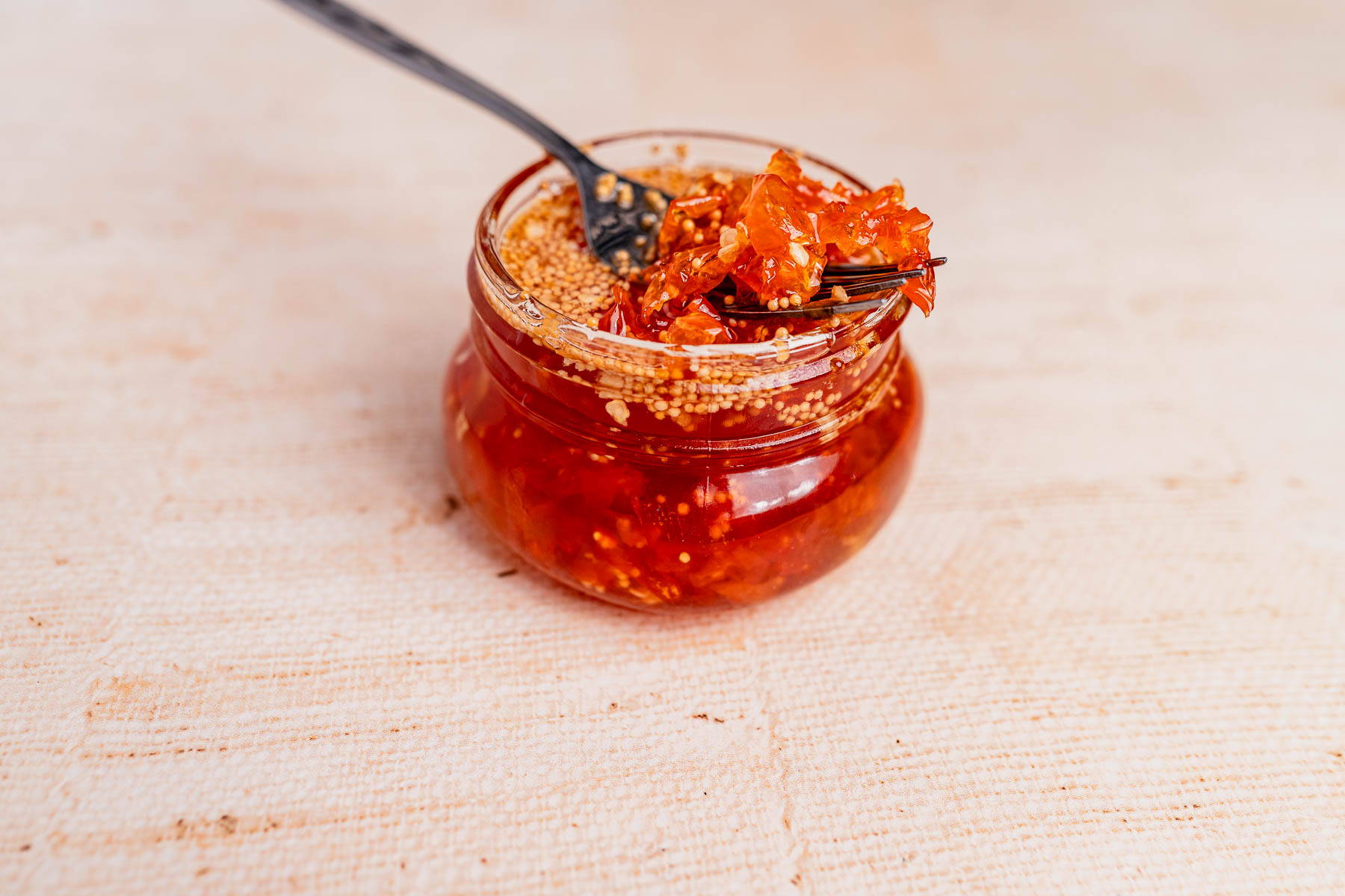 A glass jar filled with red chili sauce containing visible seeds and candied habaneros, with a black fork scooping some out.