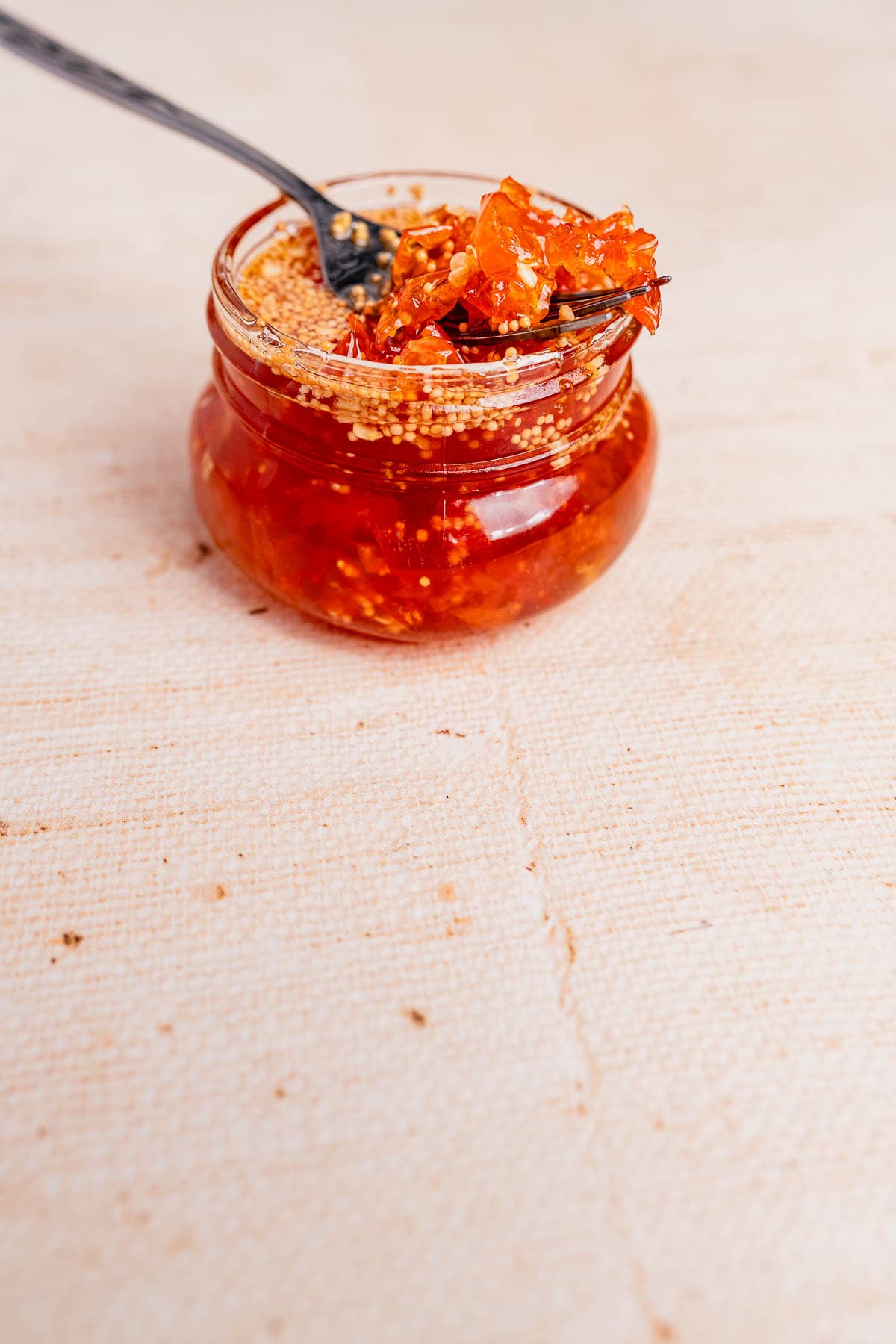 A glass jar of chili crisp with candied habaneros, with a spoon holding a portion above the open jar, placed on a light textured surface.
