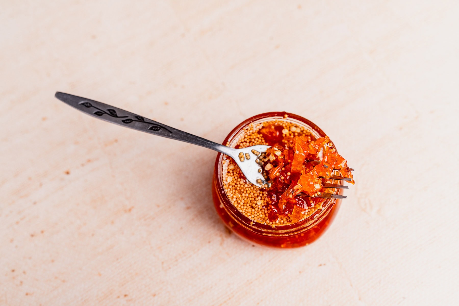 A jar of chili oil with floating seeds and candied habaneros, with a fork holding a portion of chili oil above the jar, photographed on a light surface.