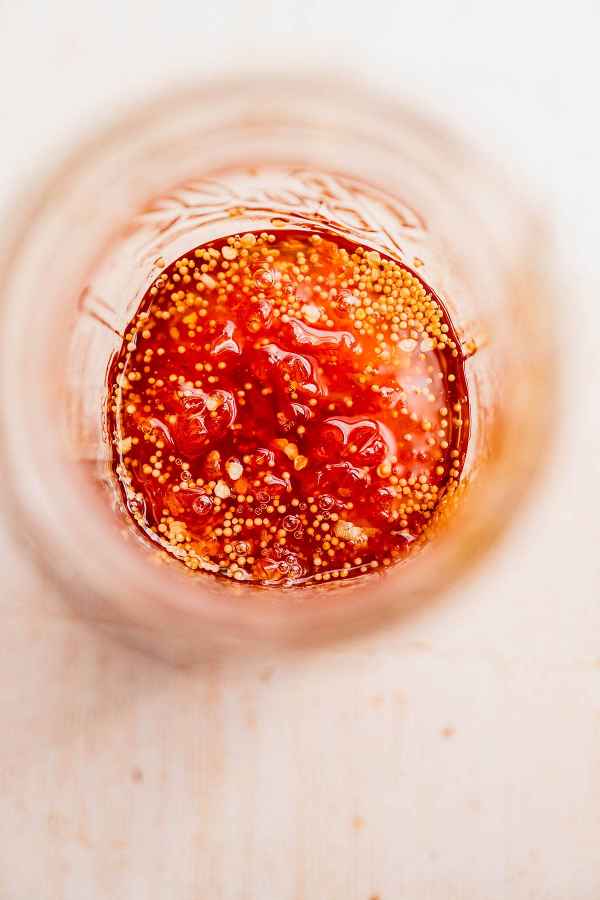 Close-up, top-down view of a glass jar containing a mixture of red sauce with visible mustard seeds and candied habaneros.
