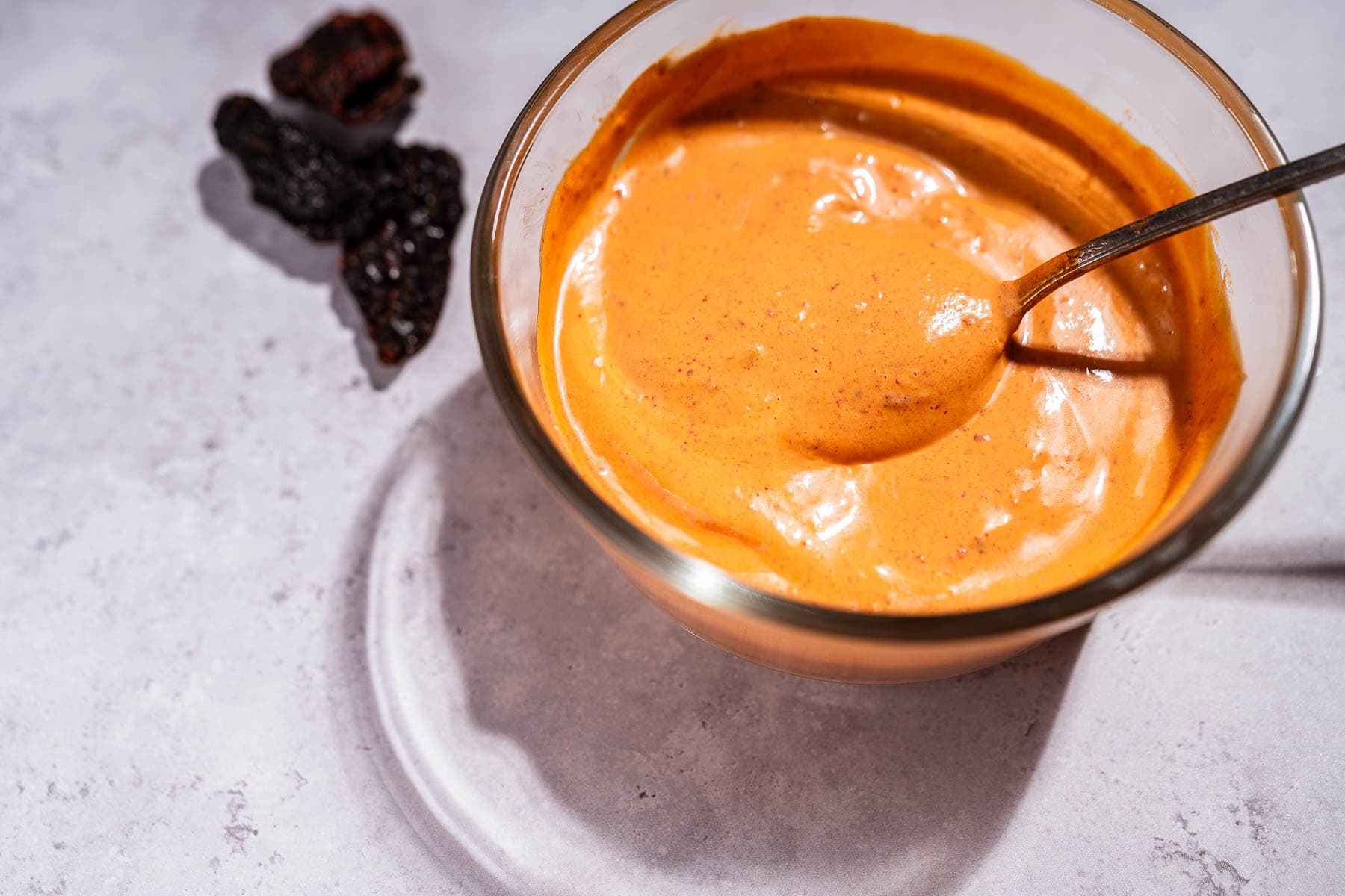 A glass bowl filled with creamy chipotle sauce, with a spoon inside, sits on a light gray surface next to three dried black chilies.