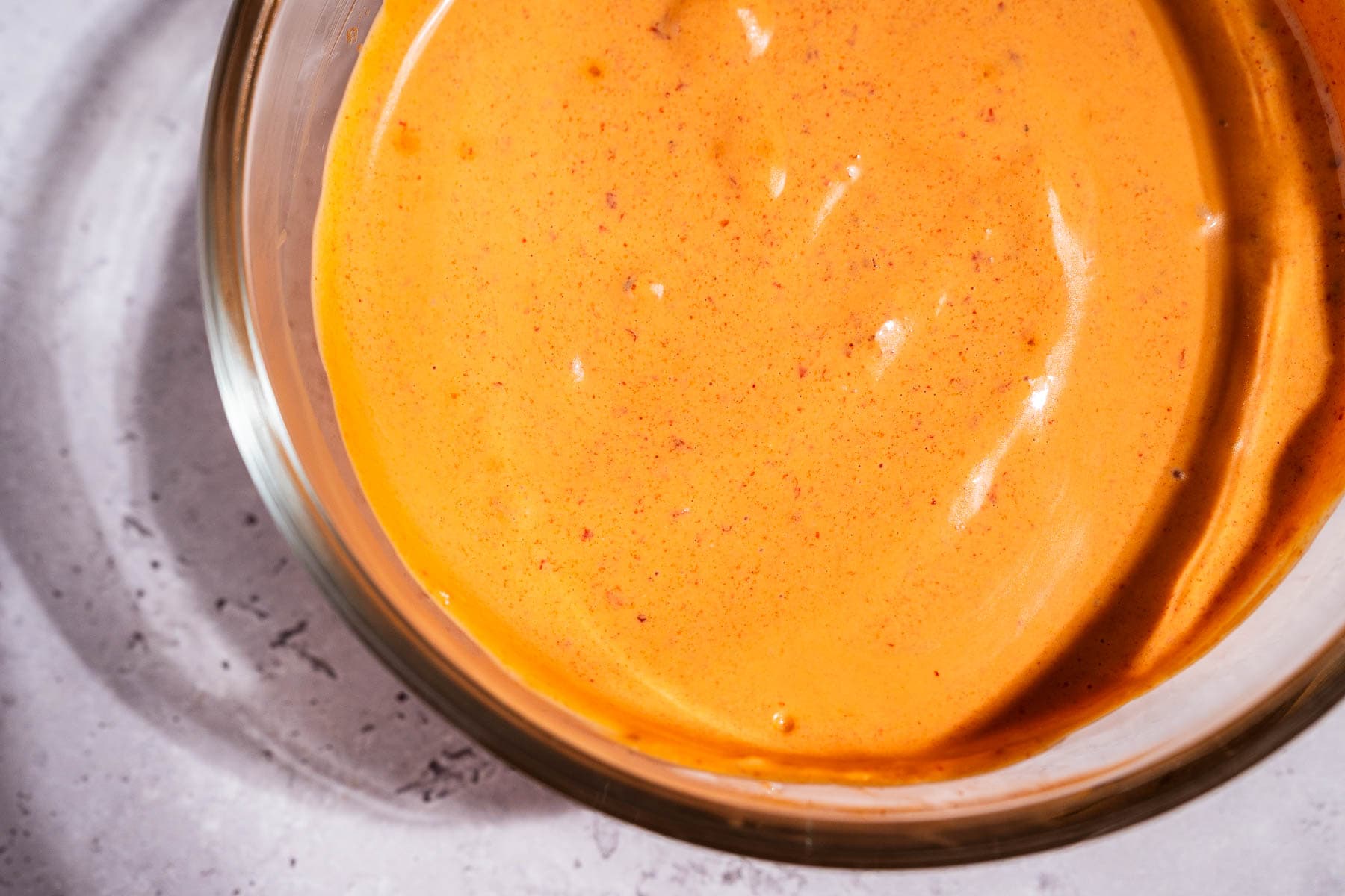 A clear glass bowl filled with creamy, orange-colored chipotle sauce sits on a light gray surface.