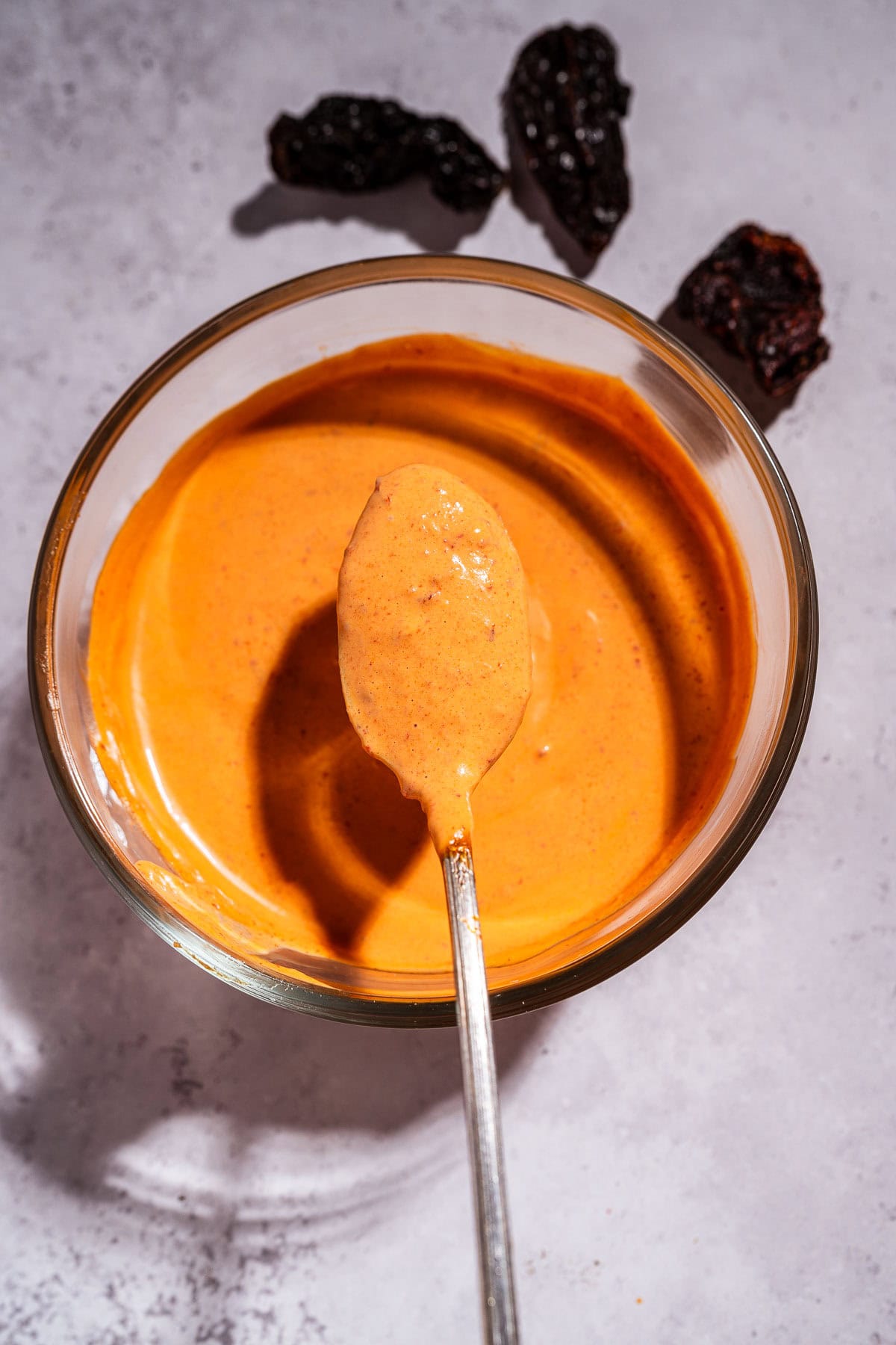 A bowl of creamy chipotle sauce with a spoon resting in it; three dried chili peppers are placed nearby on the gray surface.
