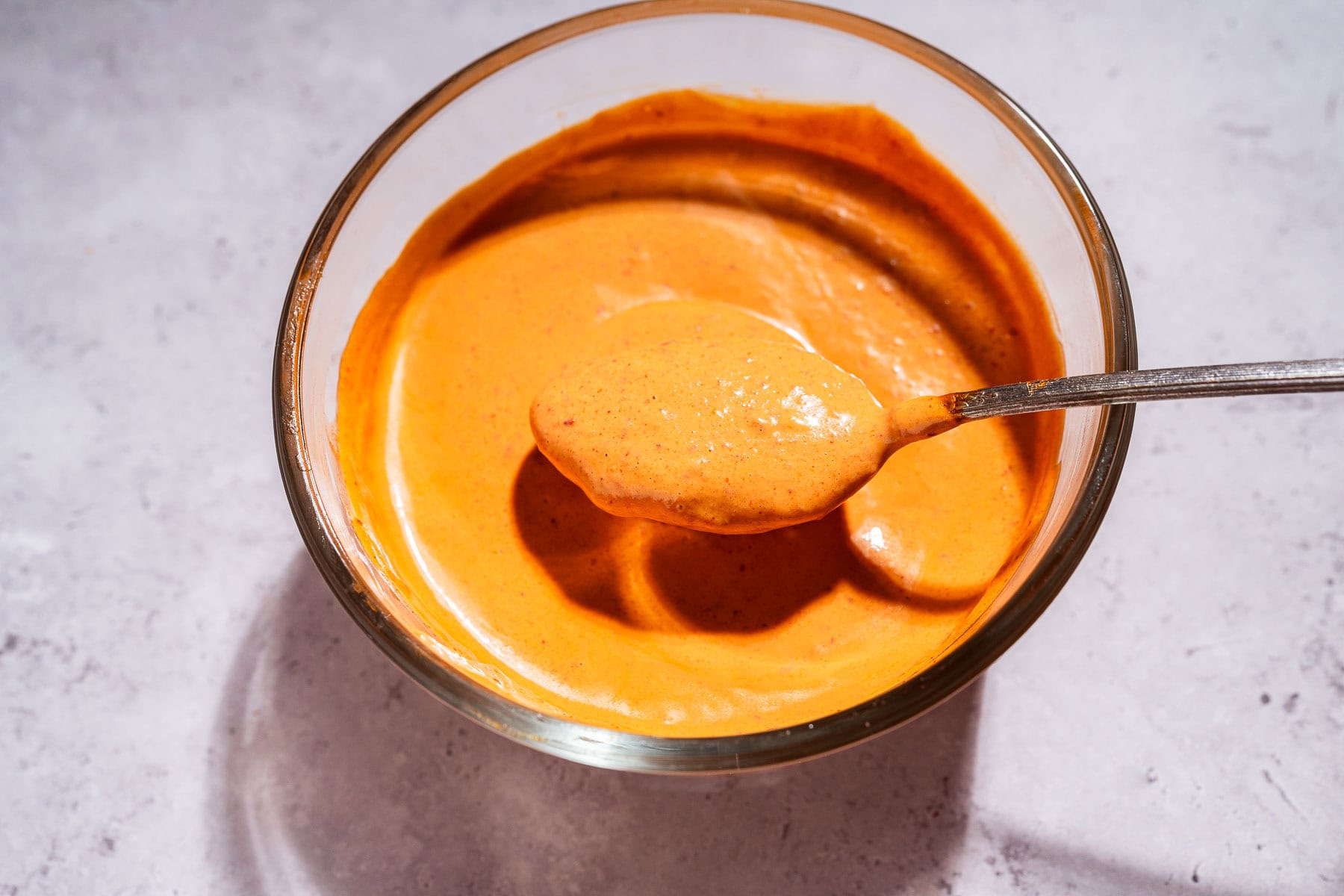 A glass bowl filled with vibrant orange chipotle sauce, with a metal spoon lifting a portion above the bowl, sits on a light textured surface.