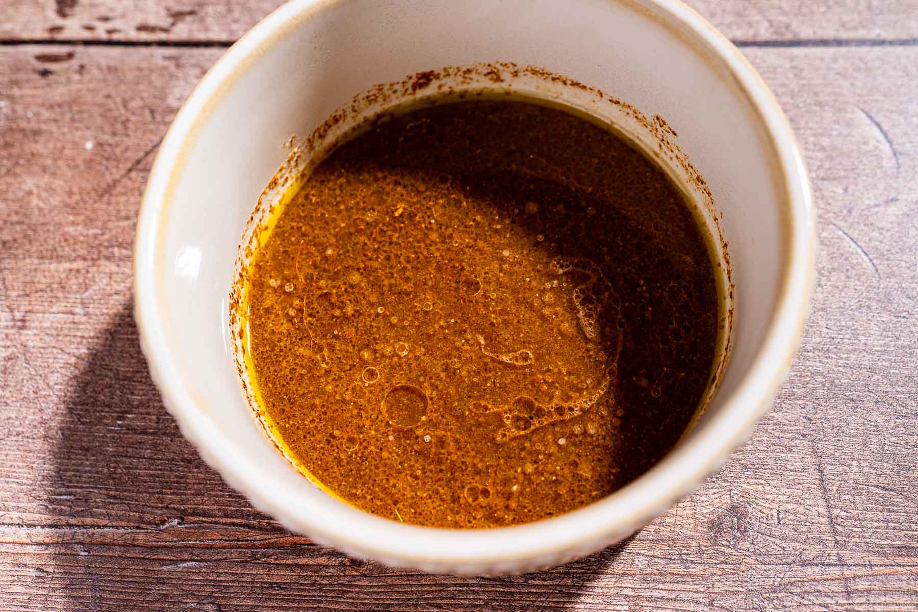 A white bowl containing a mixture of brown liquid with visible oil separation, resembling a tangy fiesta salad dressing, is placed on a wooden surface.