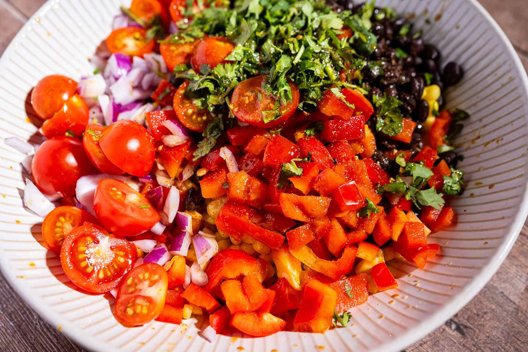 A bowl filled with chopped red bell peppers, cherry tomatoes, red onions, black beans, cilantro, and corn makes a vibrant fiesta salad.