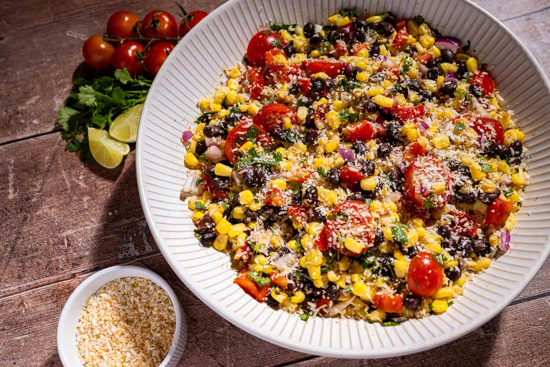 A bowl of fiesta salad with corn, black beans, tomato, red onion, and cilantro topped with shredded cheese, served alongside extra shredded cheese and tomatoes with lime.