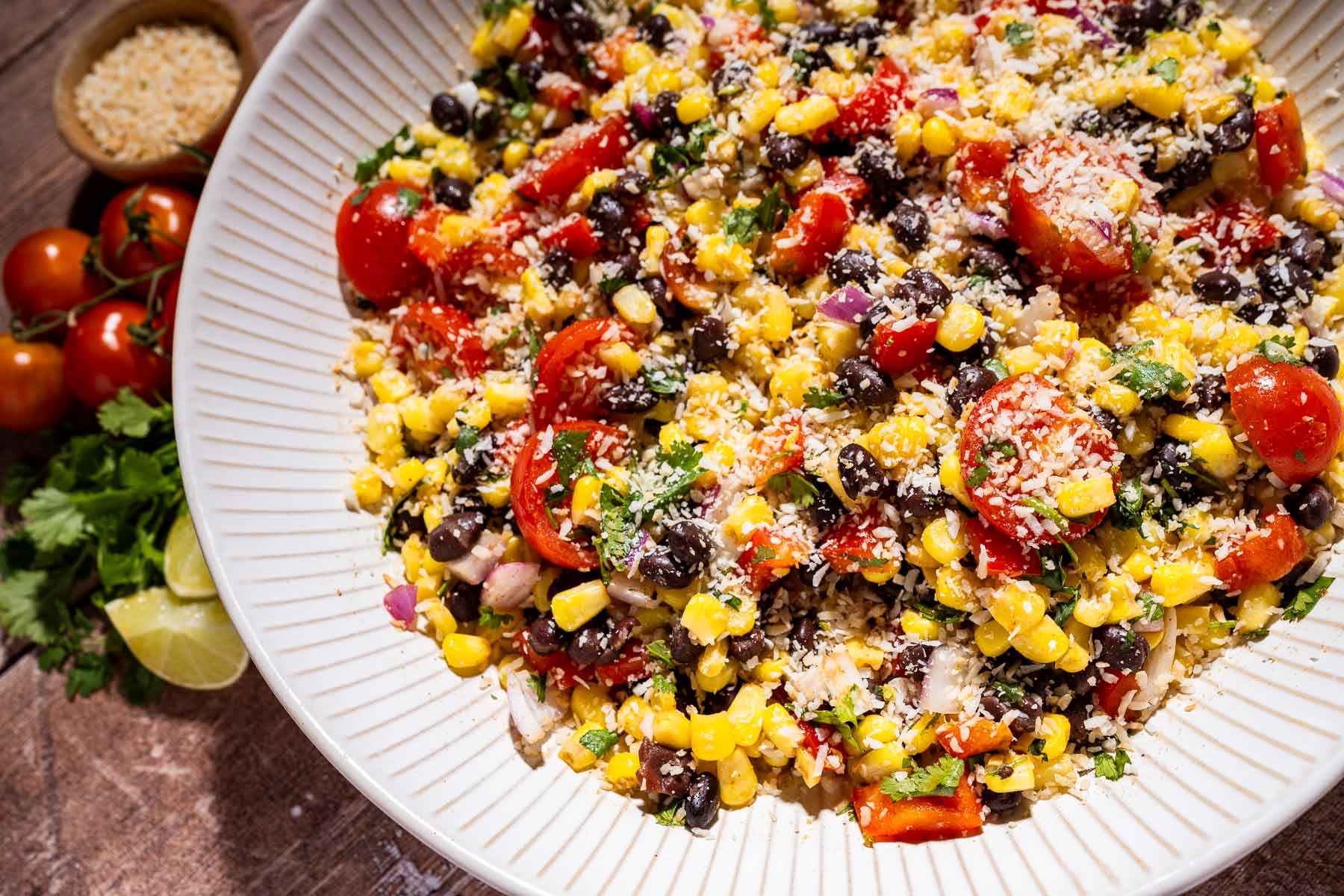 A vibrant fiesta salad with corn, black beans, cherry tomatoes, red onions, cilantro, and shredded cheese is served on a plate, with limes, tomatoes, and parmesan in the background.