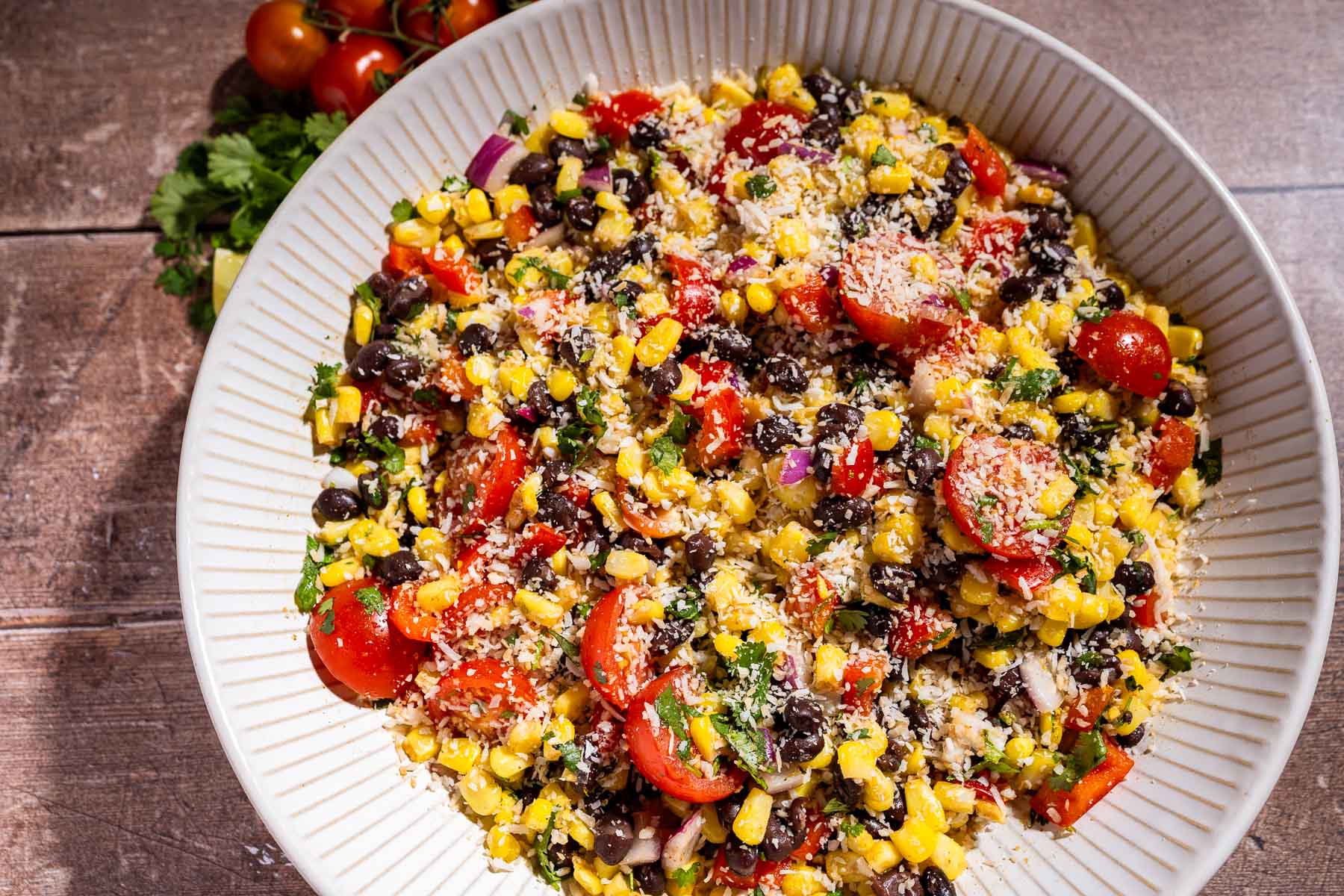 A bowl of colorful fiesta salad with corn, black beans, cherry tomatoes, red onion, cilantro, and grated cheese on top, set on a wooden table.