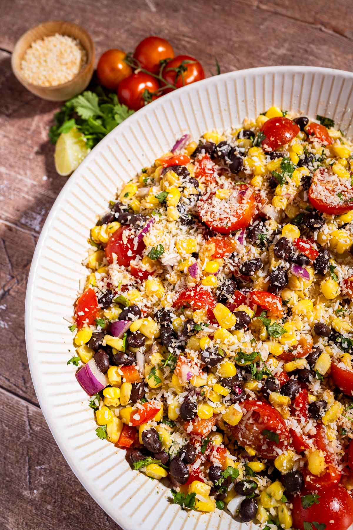 A bowl of fiesta salad with corn, black beans, cherry tomatoes, red onion, cilantro, and shredded cheese on top, set on a wooden table with tomatoes and a bowl of grated cheese nearby.