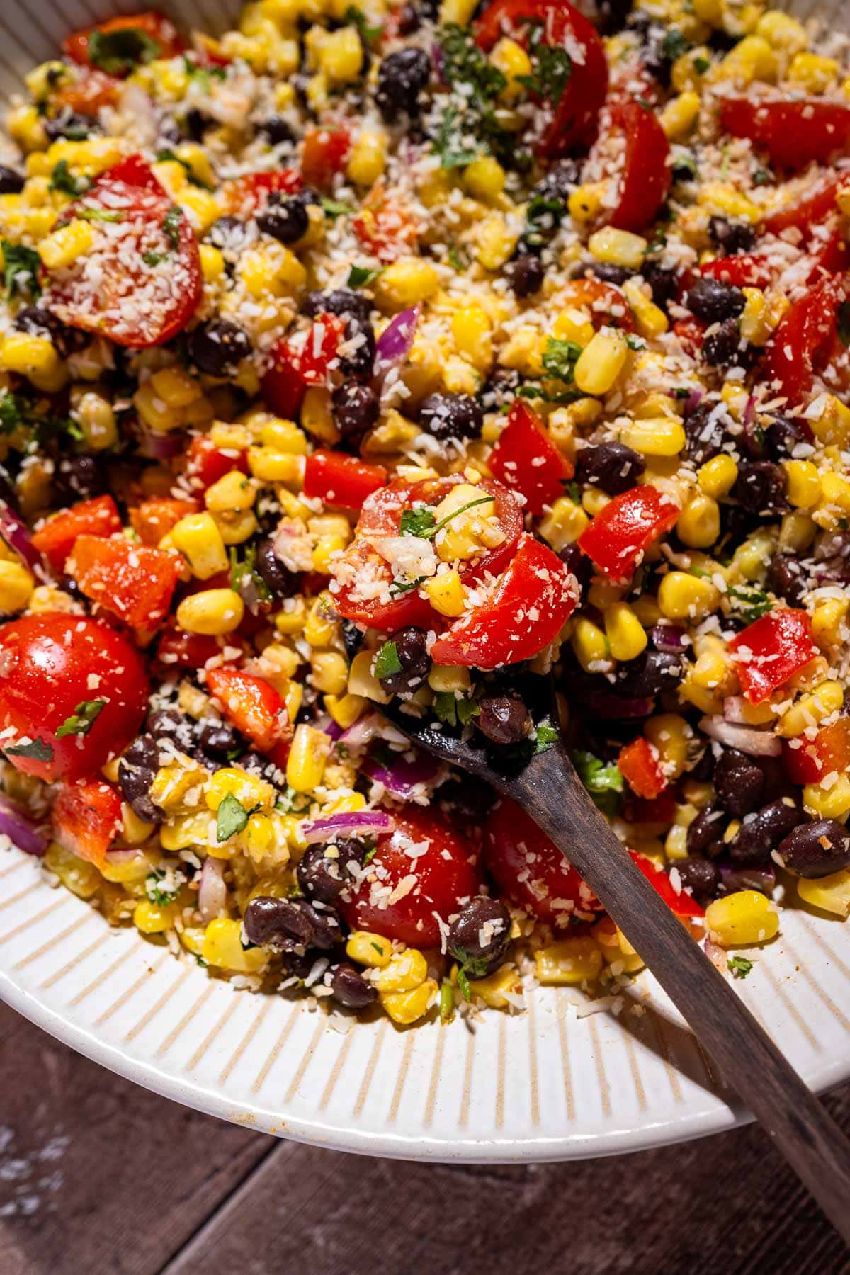 A bowl of fiesta salad featuring corn, black beans, and tomatoes mixed with chopped red onion, herbs, and grated cheese, served with a spoon.