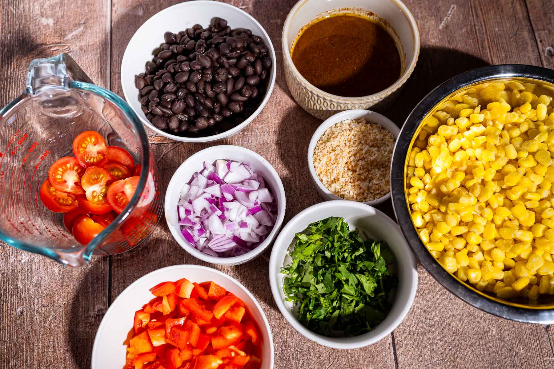 Several bowls and containers on a wooden surface, each holding fiesta salad ingredients: cherry tomatoes, black beans, chili sauce, corn, diced onion, breadcrumbs, chopped cilantro, and red bell pepper.