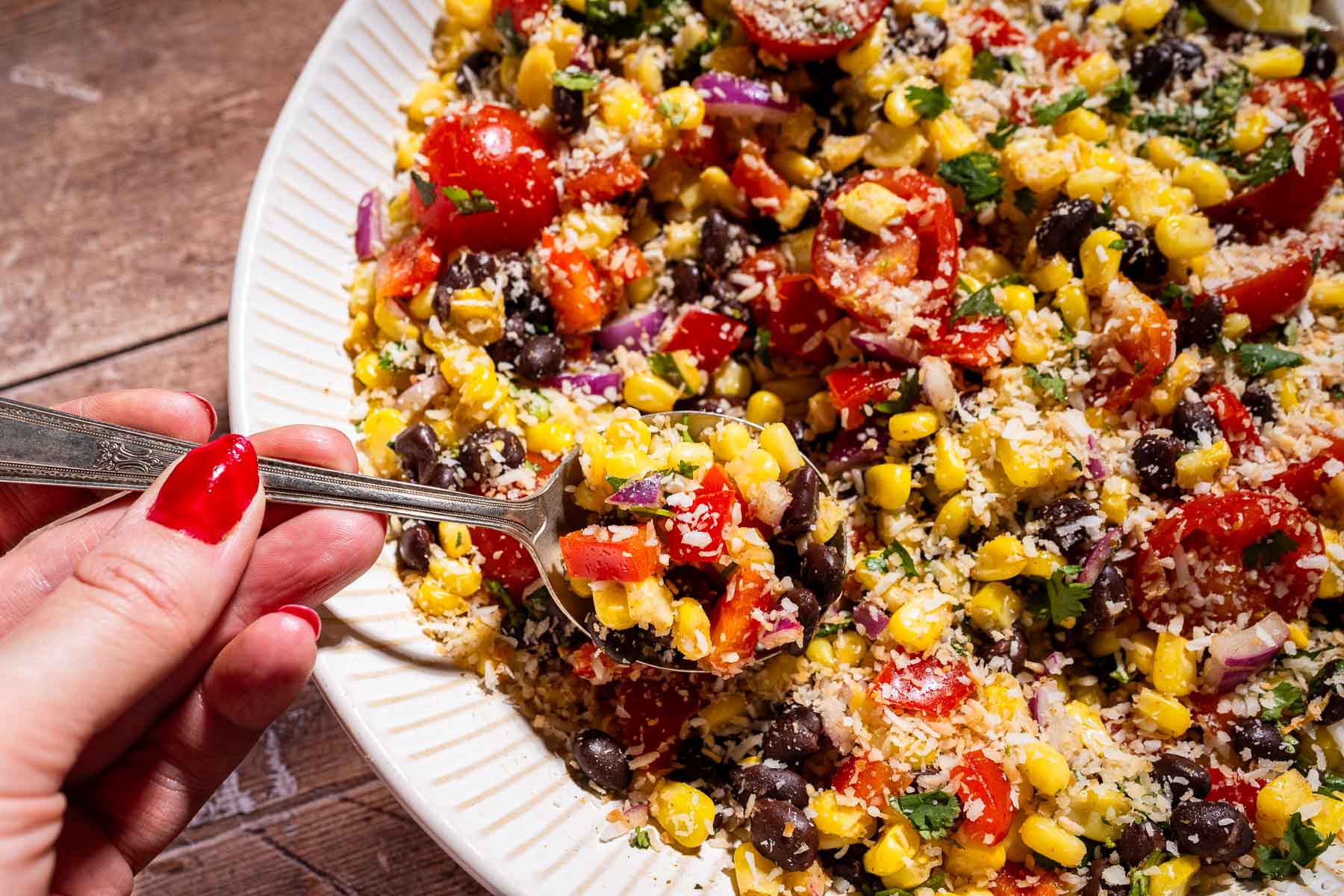 A hand holding a spoon scoops a vibrant fiesta salad with corn, black beans, cherry tomatoes, red onions, red peppers, and grated cheese from a white plate.