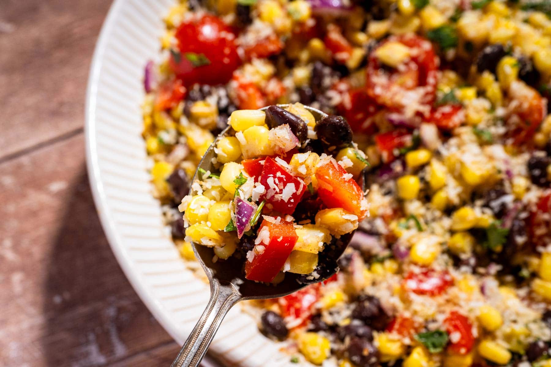 A close-up of a spoon holding fiesta salad with corn, black beans, chopped tomatoes, red onions, and shredded cheese over a plate of the same colorful salad.