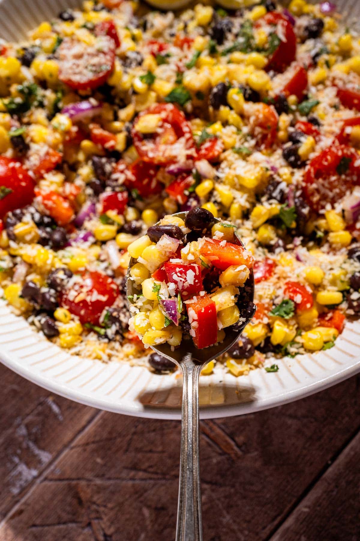 A spoonful of fiesta salad—corn, black bean, red onion, tomato, and cilantro—hovers above a bowl of the same fresh mixture, all topped with grated cheese.