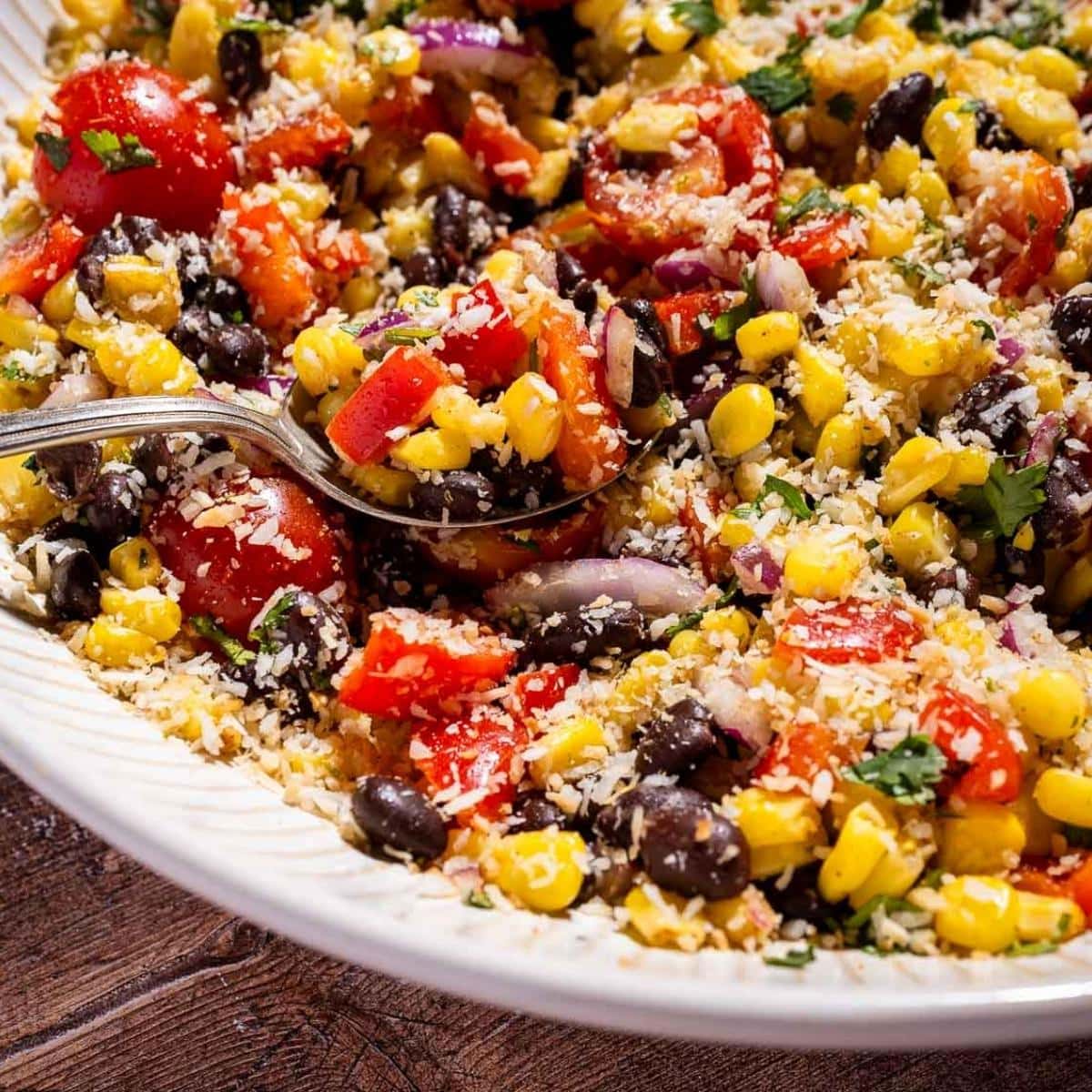 A close-up of a fiesta salad with corn, black beans, red peppers, tomatoes, red onions, herbs, and shredded cheese in a white bowl with a spoon.