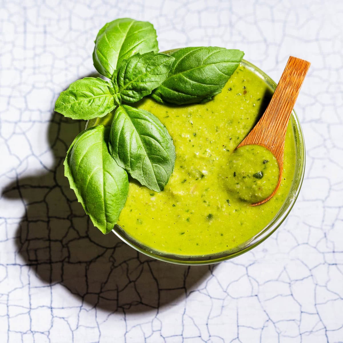 A bowl of green pesto salad dressing with a wooden spoon and fresh basil leaves on top, placed on a white cracked surface.