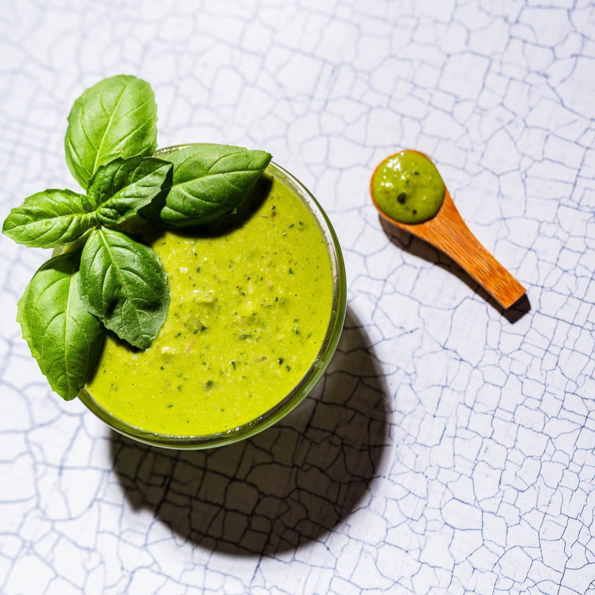 A glass bowl of green pesto salad dressing garnished with fresh basil leaves, with a small wooden spoon containing pesto on a white textured surface.