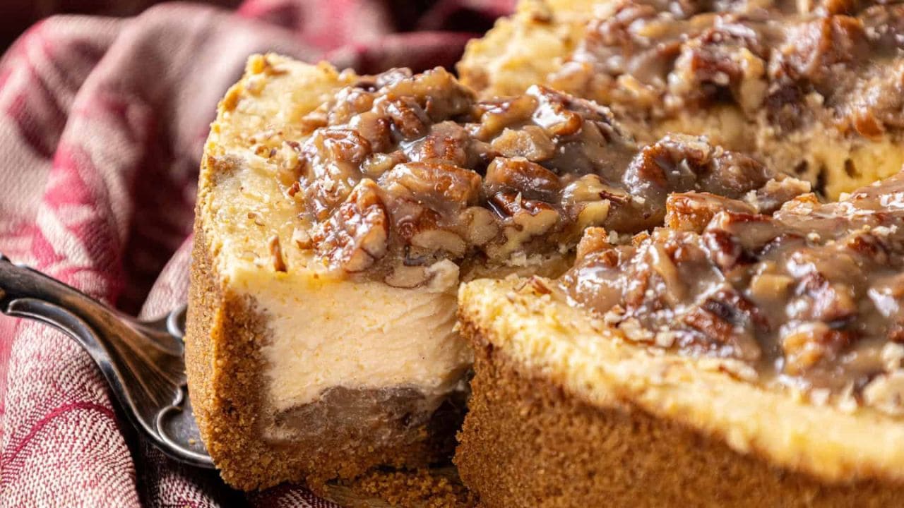 The image features an Apple Bottom Bourbon Pecan Cheesecake with chopped pecans and a caramel glaze. A slice is slightly separated from the whole, revealing the rich filling and thick graham cracker crust.
