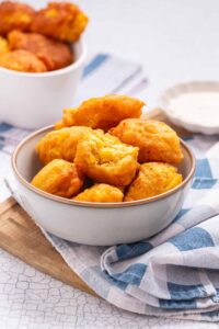 A gray bowl filled with golden, crispy hush puppies sits on a striped cloth napkin, with more hush puppies in a white bowl in the background.