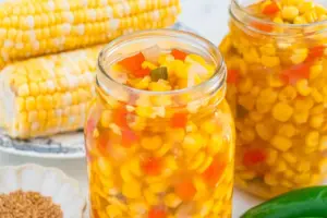 Jars filled with corn relish containing corn kernels, diced red peppers, and onions, with whole corn cobs and spices in the background.