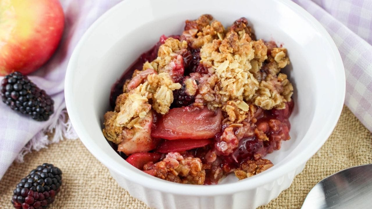 A bowl of fruit crumble with oats on top, featuring apples and blackberries. A fresh apple and blackberries are placed beside the bowl.