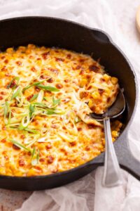 A skillet of melted cheesy corn topped with sliced green onions, with a serving spoon scooping some out.