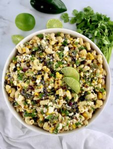 A bowl of pasta salad with corn, black beans, red onion, cilantro, crumbled cheese, and lime slices, placed on a white surface with fresh cilantro, lime, and a pepper nearby.