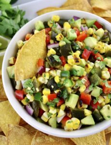 A bowl of colorful corn salsa with diced avocado, red pepper, onion, cilantro, and tortilla chips on the side.