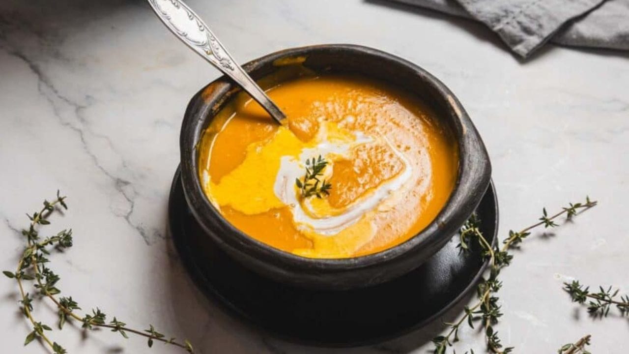 A bowl of creamy orange soup is placed on a black plate, garnished with a swirl of cream and a sprig of herb. A spoon rests inside the bowl. Small sprigs of thyme are scattered on the marble surface nearby.