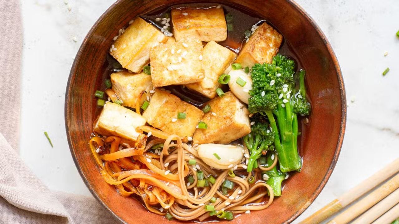 A bowl of soba noodles in broth, topped with fried tofu, broccolini, carrot spirals, and sliced mushrooms. Chopsticks rest on the bowl, and sesame seeds are sprinkled over the dish.