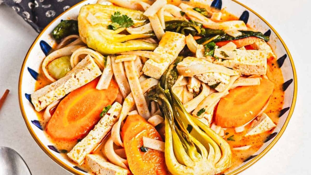 A bowl of noodle soup with tofu strips, baby bok choy, carrot slices, and fresh herbs, set on a white surface with a spoon nearby and a small bowl of chopped greens in the background.