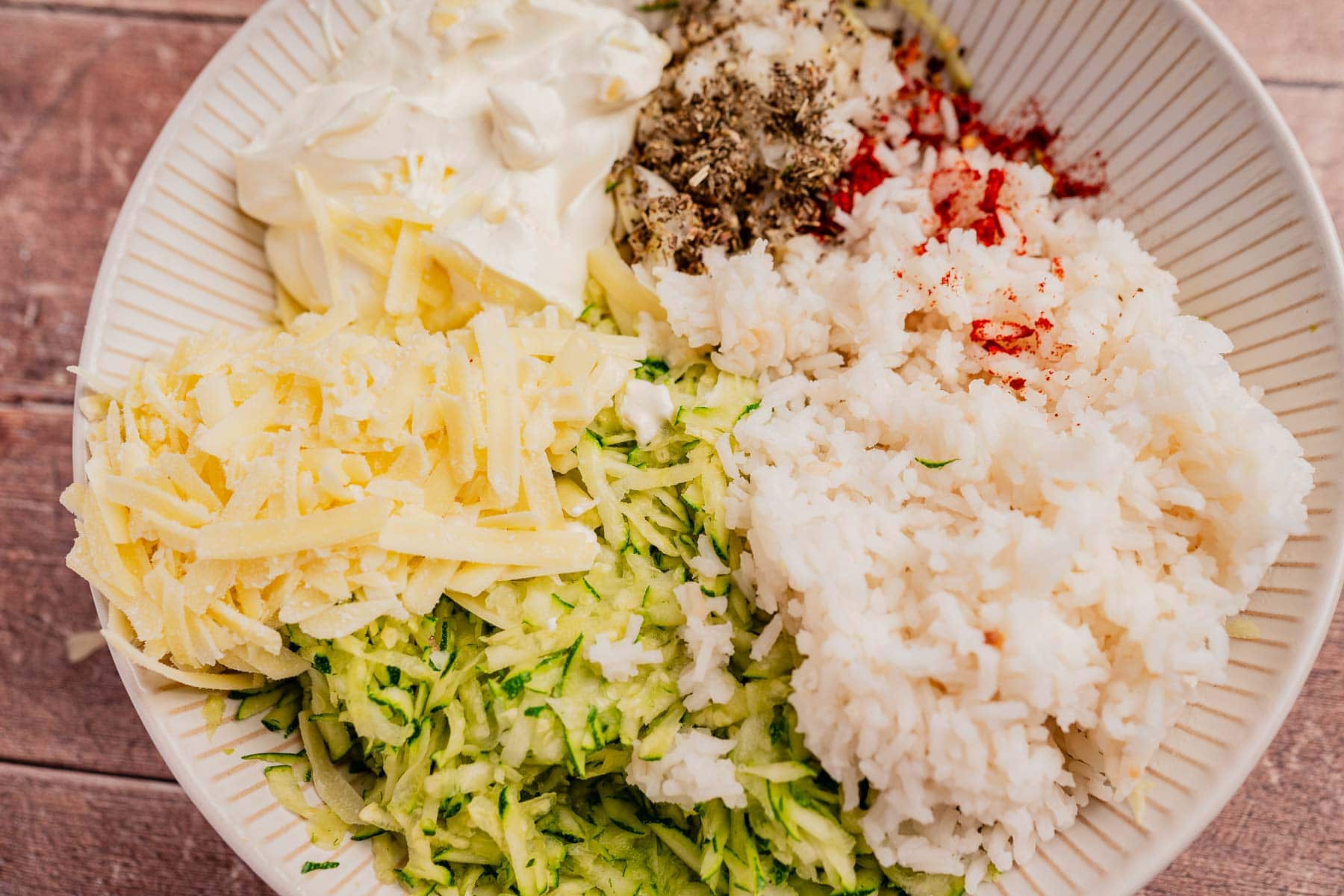 A bowl containing ingredients for zucchini rice casserole—grated zucchini, cooked rice, shredded cheese, mayonnaise, and assorted spices—arranged in separate sections.