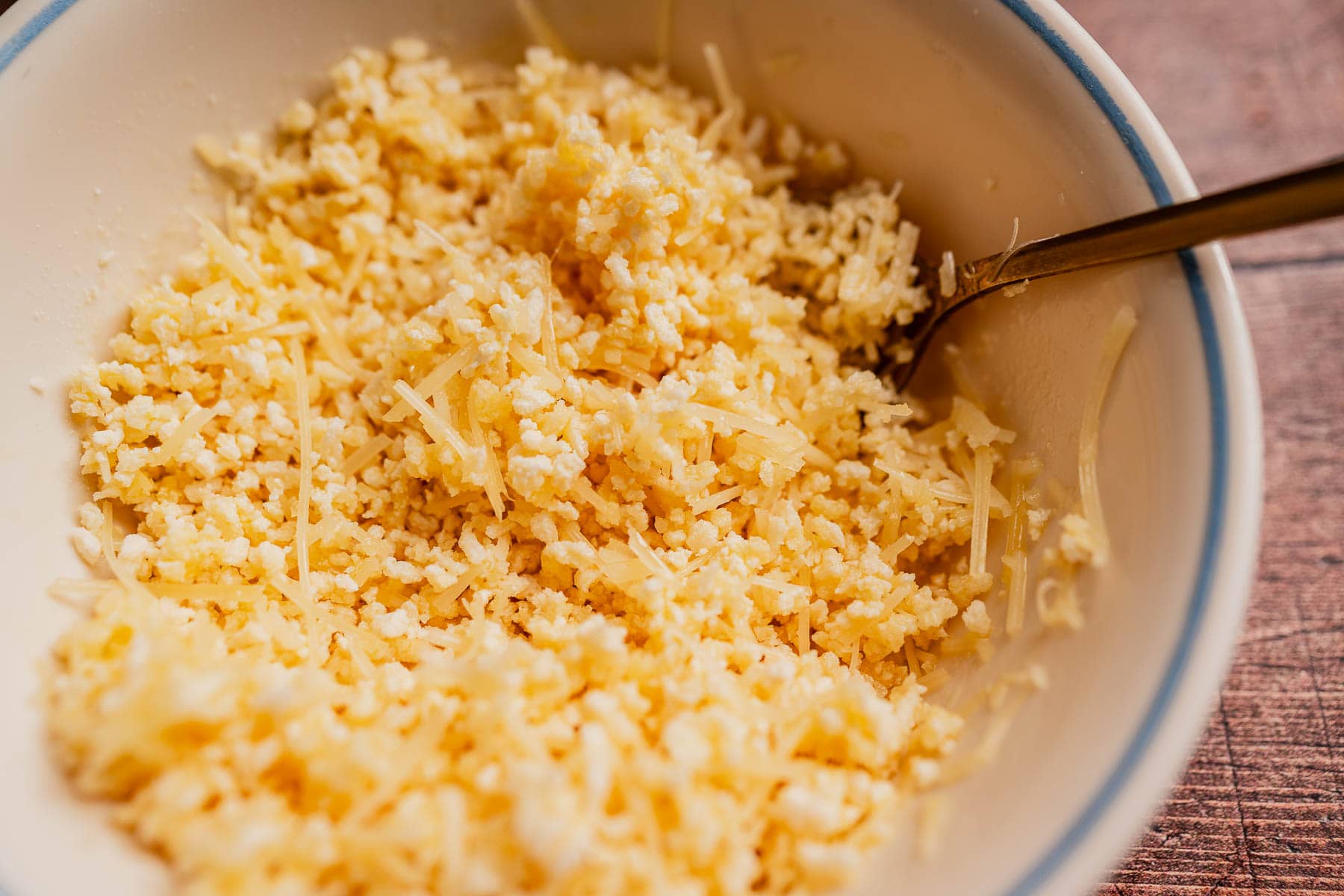 A bowl filled with shredded and grated cheese, perfect for sprinkling over a zucchini rice casserole, with a metal spoon resting on the side.
